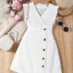 Vestido blanco para niñas con botones decorativos y volantes, exhibido en percha con accesorios de verano.