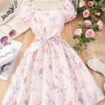 Vestido rosa floral con manga farol y cinturón en percha de madera, decorado con rosas alrededor.